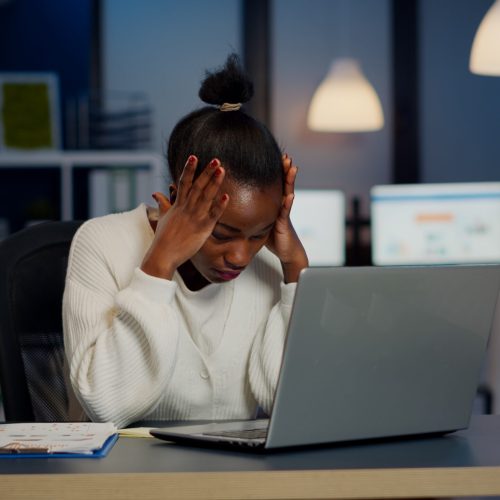 Stressed african businesswoman suffering from headache at work doing overtime late night. Exhausted tensed worker coping with migraine, emotional stress concept feeling pain working at laptop